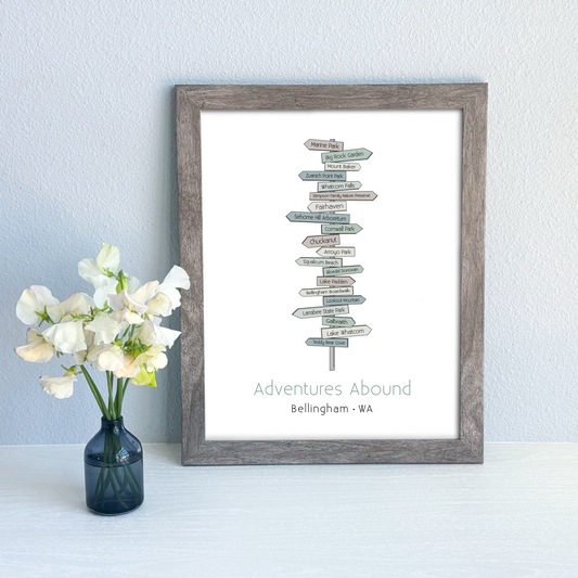 Framed artwork with 'Adventures Abound' text and a vase of flowers on a light blue wall.