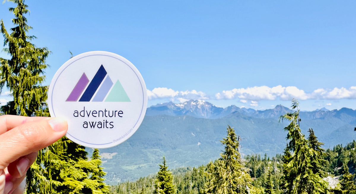 Hand holding up a sticker in front of a mountain-scape. The sticker has the words "adventure awaits" underneath a colorful rendering of mountain peaks