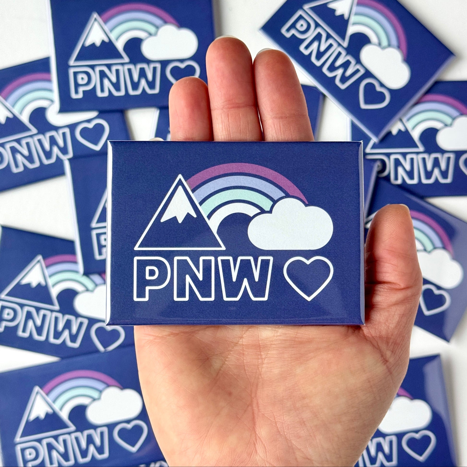 Hand holding a blue magnet with PNW design surrounded by more magnets on a white background