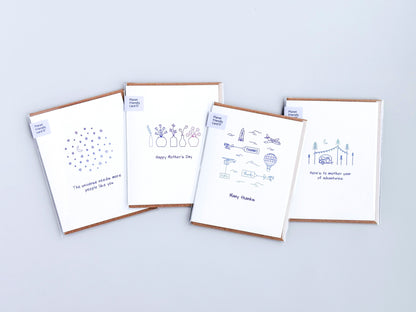 Four white greeting cards with kraft brown envelopes, arranged in a fan shape on a light gray background. Each card features a simple line drawing and a short message, including 'The universe needs more people like you', 'Happy Mother's Day', 'Many thanks', and 'Here's to another year of adventures'.