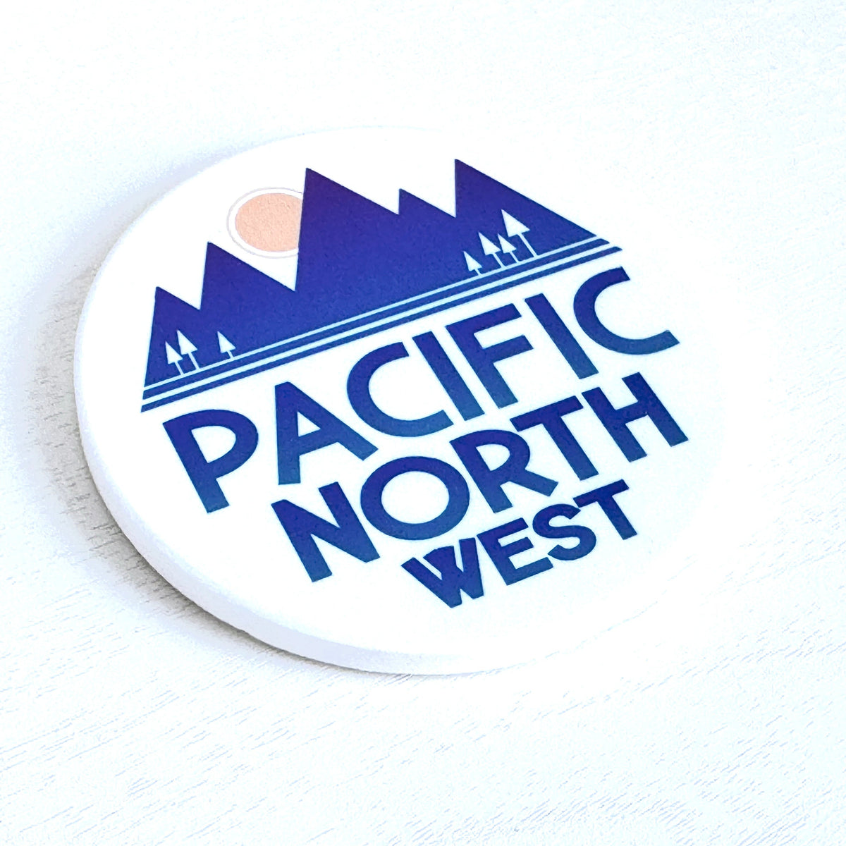 A round white coaster featuring a blue graphic of mountains, trees, and a sun with the text 'PACIFIC NORTH WEST' in blue, shown on a light textured background.