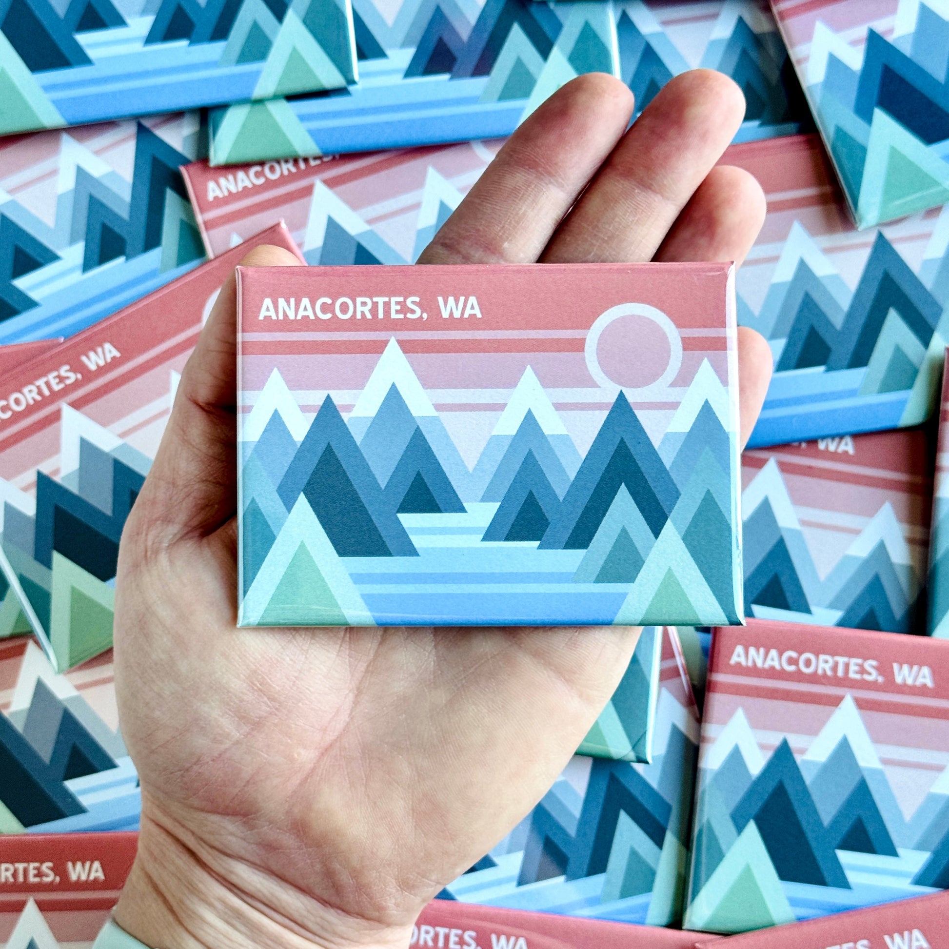 Hand holding a fridge magnet with mountain design and 'Anacortes, WA' text among more magnets.