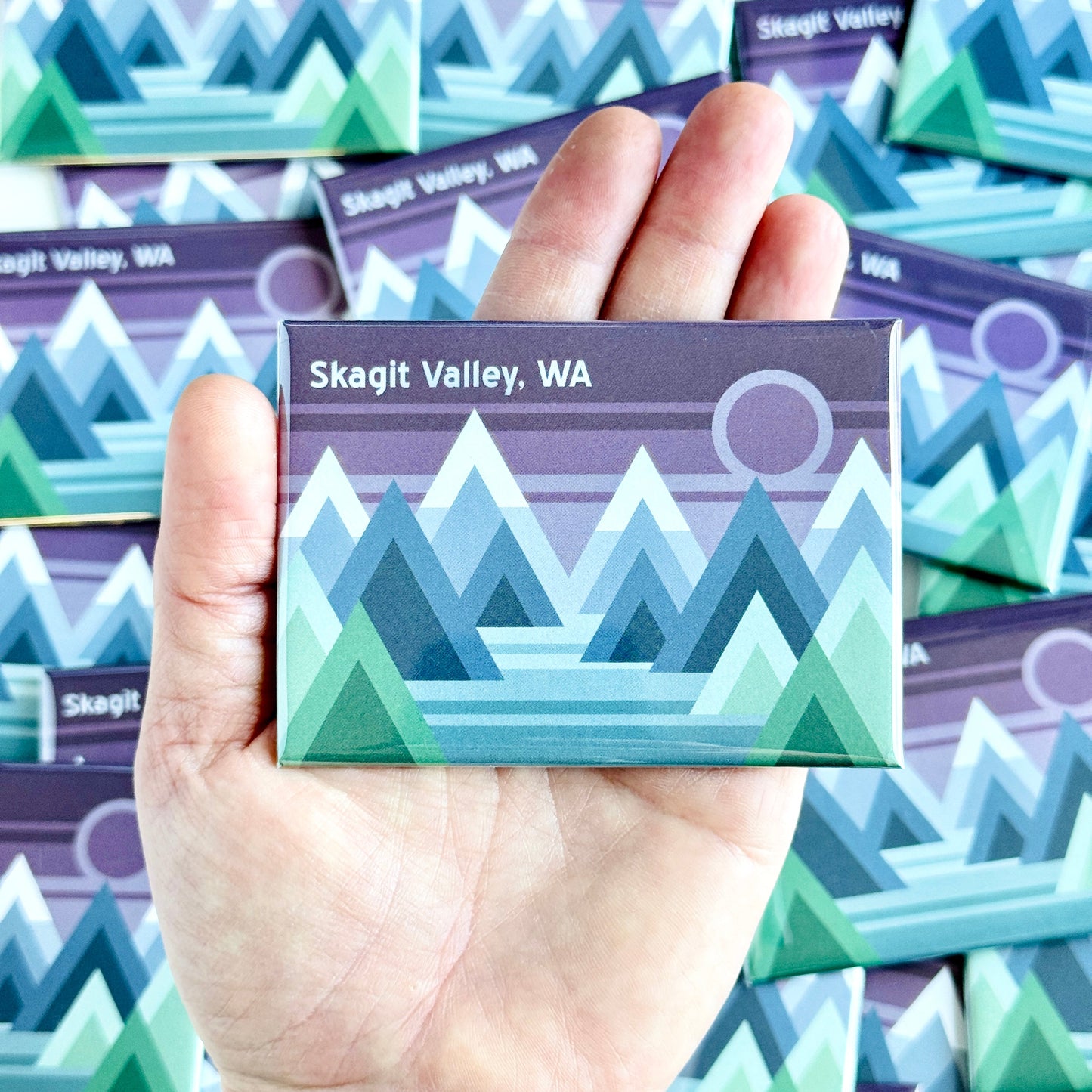 Hand holding a magnet with mountain design and 'Skagit Valley, WA' text against a background of similar magnets.