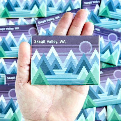Hand holding a magnet with mountain design and 'Skagit Valley, WA' text against a background of similar magnets.