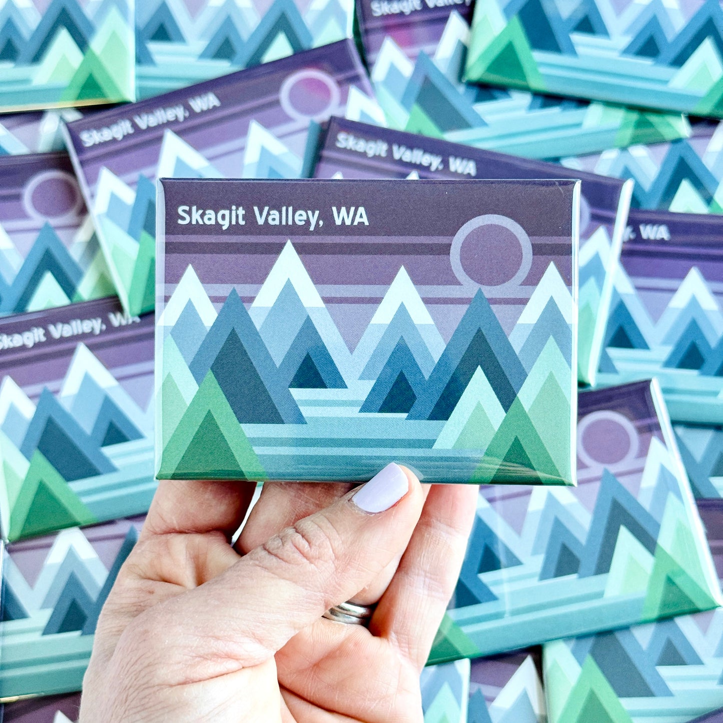 Hand holding a Skagit Valley, WA magnet with mountain design against a background of similar magnets.