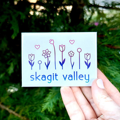 Hand holding a card with floral designs and 'Skagit Valley' text against a green forest background